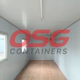 Interior of an empty shipping container with a window and wood floor.
