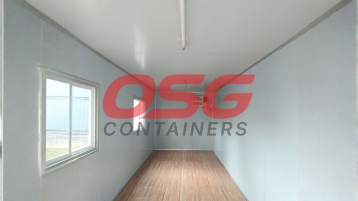 Interior of an empty shipping container with a window and wood floor.