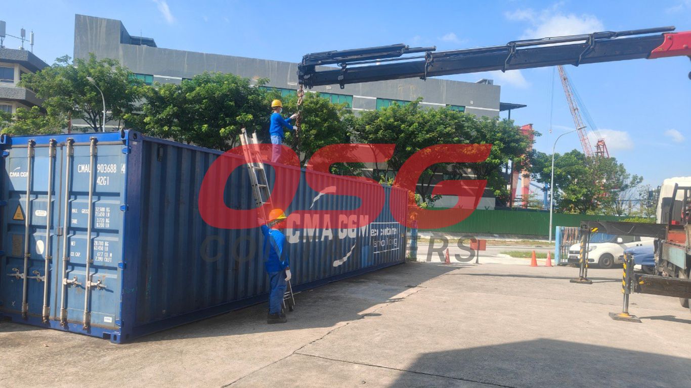 Workers use a crane and ladder to load a shipping container.