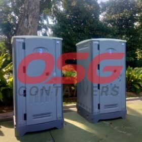 Two portable toilets are set up outdoors with lush greenery.