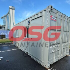 Gray shipping containers are parked in a lot with a building in the background.