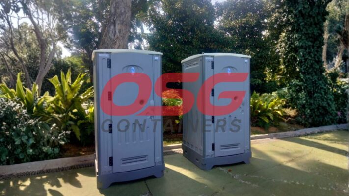 Two portable toilets are set up outdoors with lush greenery.