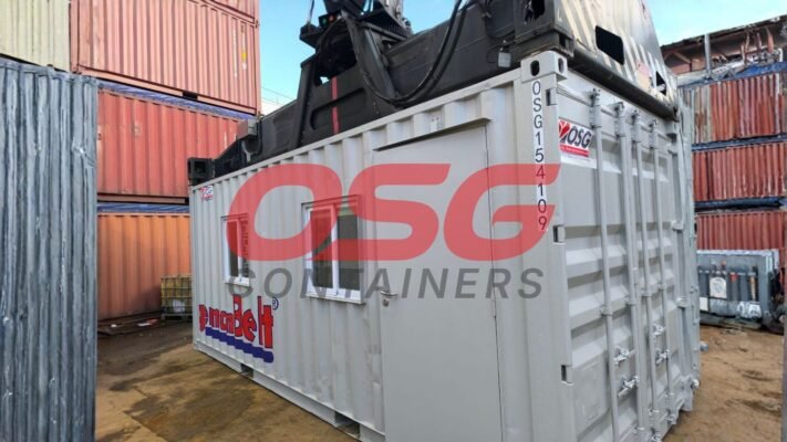 A crane lifts a modified shipping container with windows and a door.