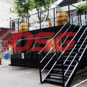 Outdoor bar setup with shipping containers, barrels, and seating.