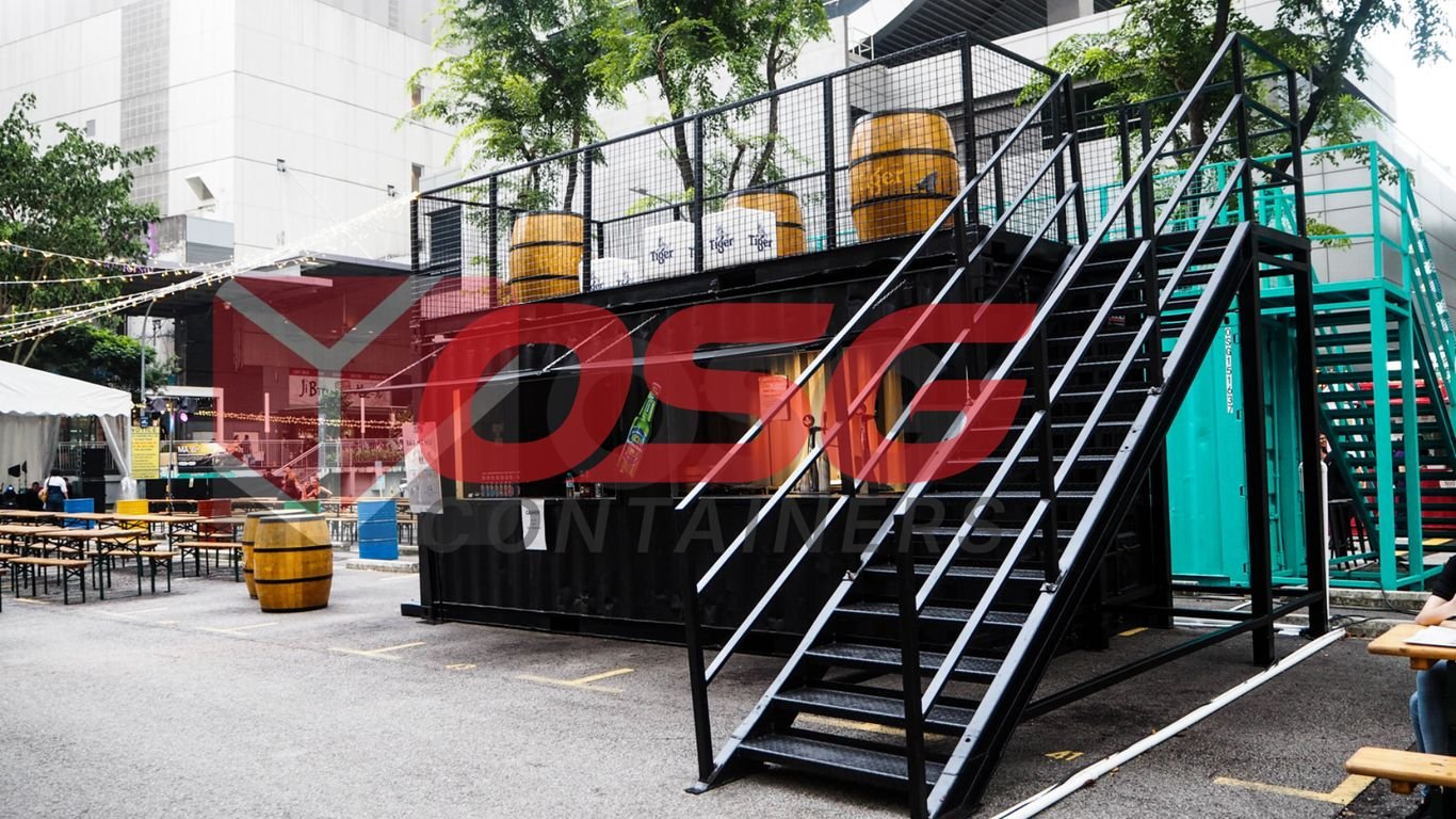 Outdoor bar setup with shipping containers, barrels, and seating.