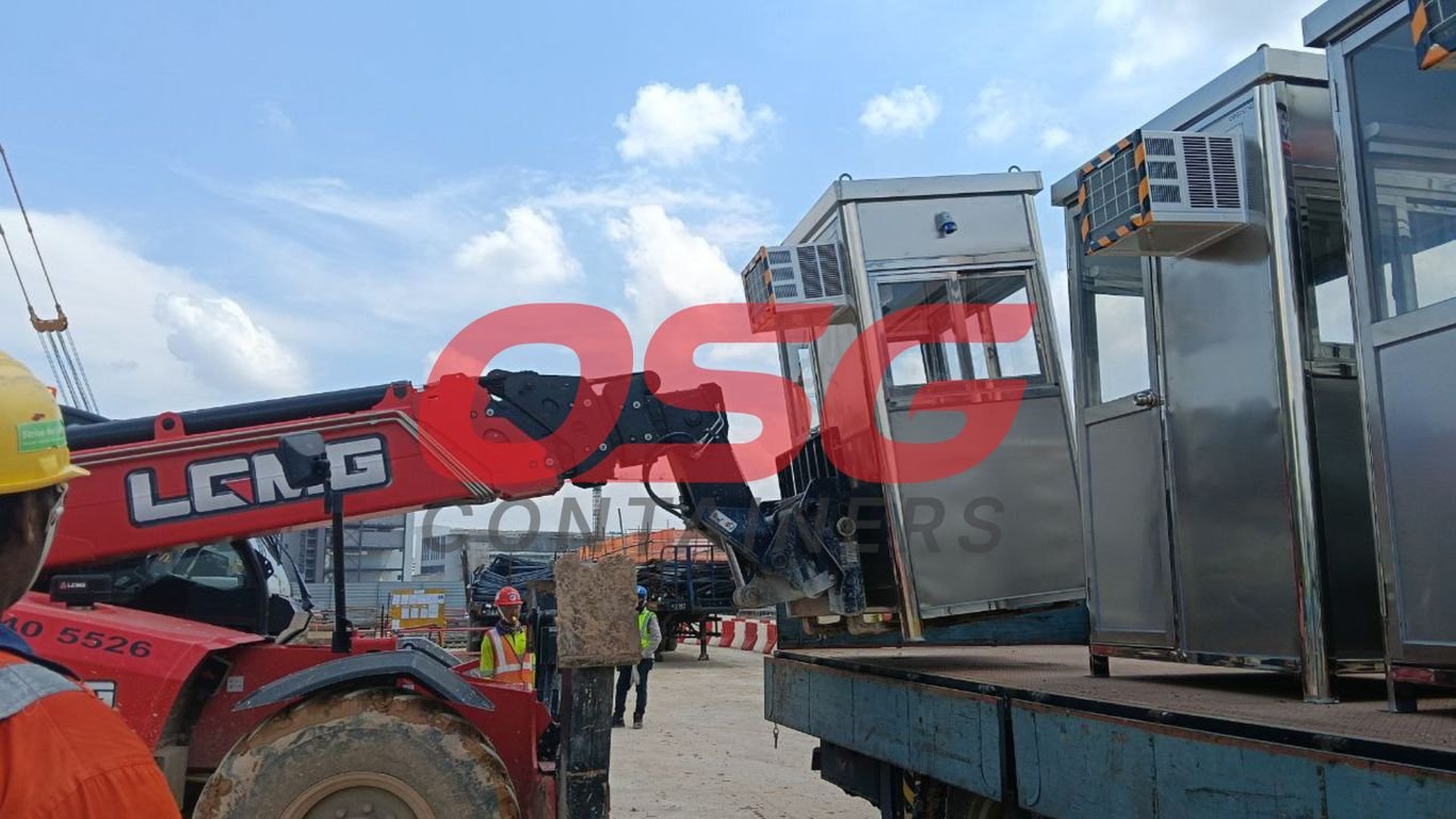 A crane lifts a security booth onto a truck at a construction site.