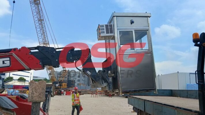 A crane lifts a portable security booth at a construction site.